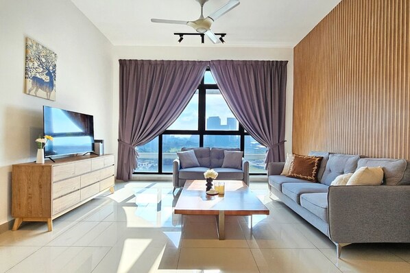 Una sala de estar moderna con un televisor de pantalla plana, un mueble de madera, un sofá gris, una mesa de centro y un ventanal con cortinas moradas.