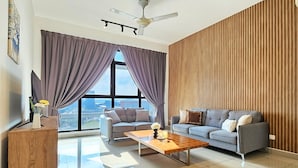 Una sala moderna con dos sofás grises, una mesa de centro de madera, un televisor sobre un mueble de madera y ventanas amplias con cortinas moradas.
