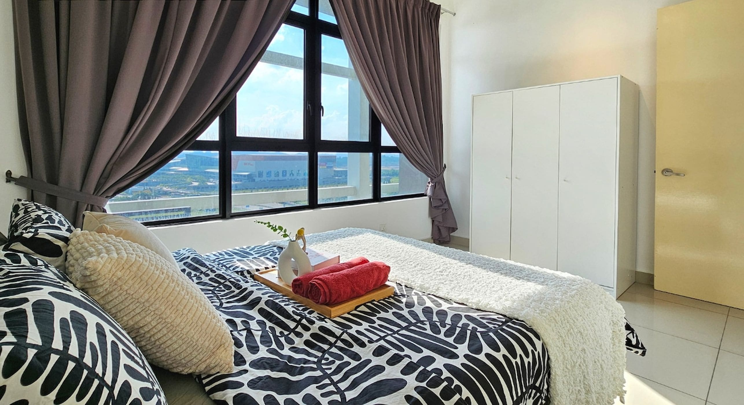A bedroom with a large window, a bed with patterned bedding, a white wardrobe, and a view of the city.