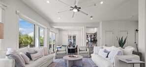 A spacious living room with a ceiling fan, large windows, and a view of palm trees. - Pelican Bluff, a premier beachside home. Private pool and elevator by Kaiser Vacations (Orange Beach)