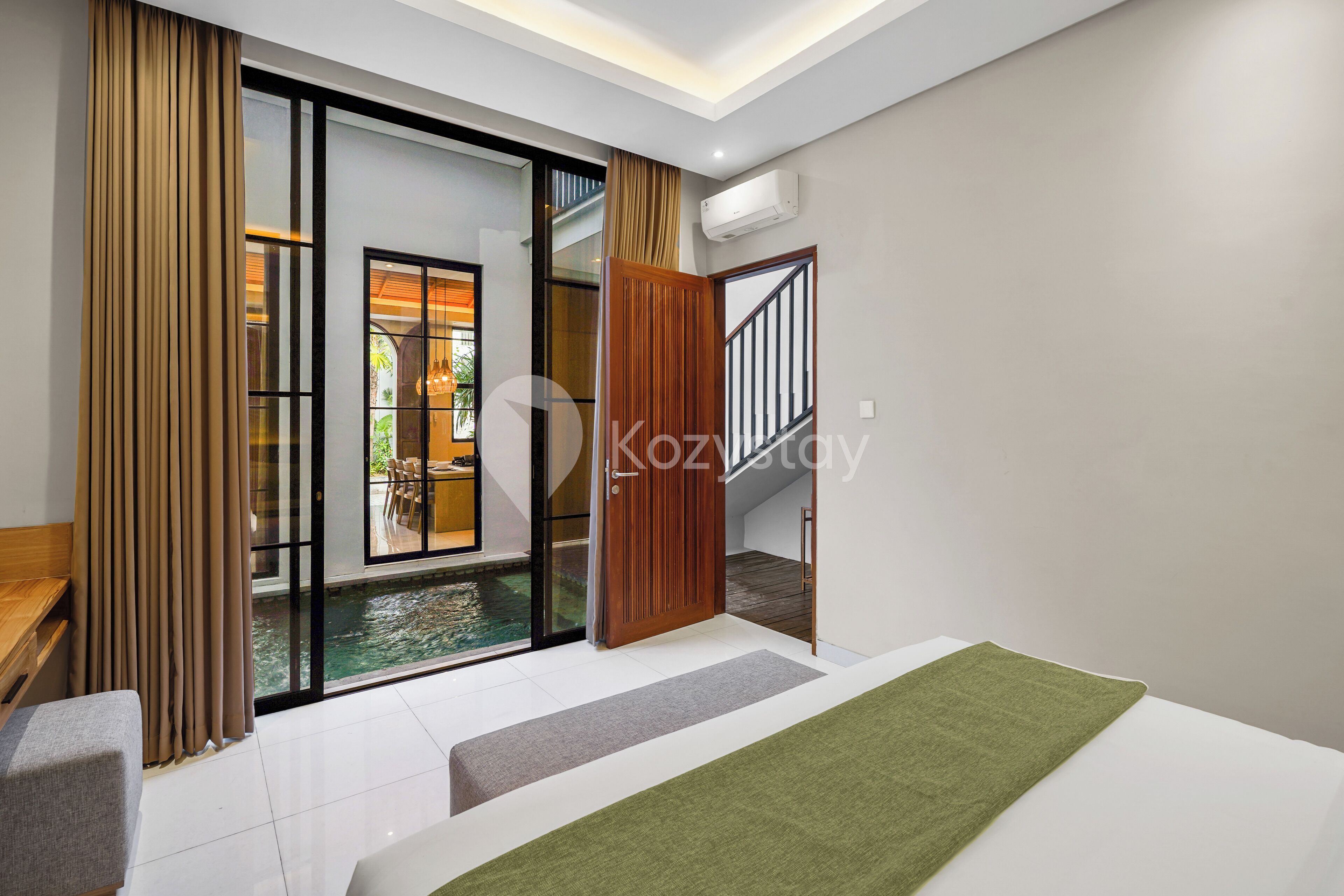 Une chambre d’hôtel moderne dotée d’une grande porte-fenêtre coulissante donnant sur un balcon avec une piscine, un banc en bois et un plaid vert sur le lit.