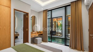 A modern hotel room with a large bed, a vanity with a mirror, a small seating area, and a view of a pool through large glass doors. - Lucenta Villa by Kozystay - Canggu (Canggu)