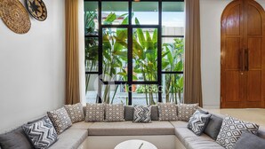 A modern living room with a large window, a sofa with patterned cushions, and a coffee table with a plant. - Lucenta Villa by Kozystay - Canggu (Canggu)