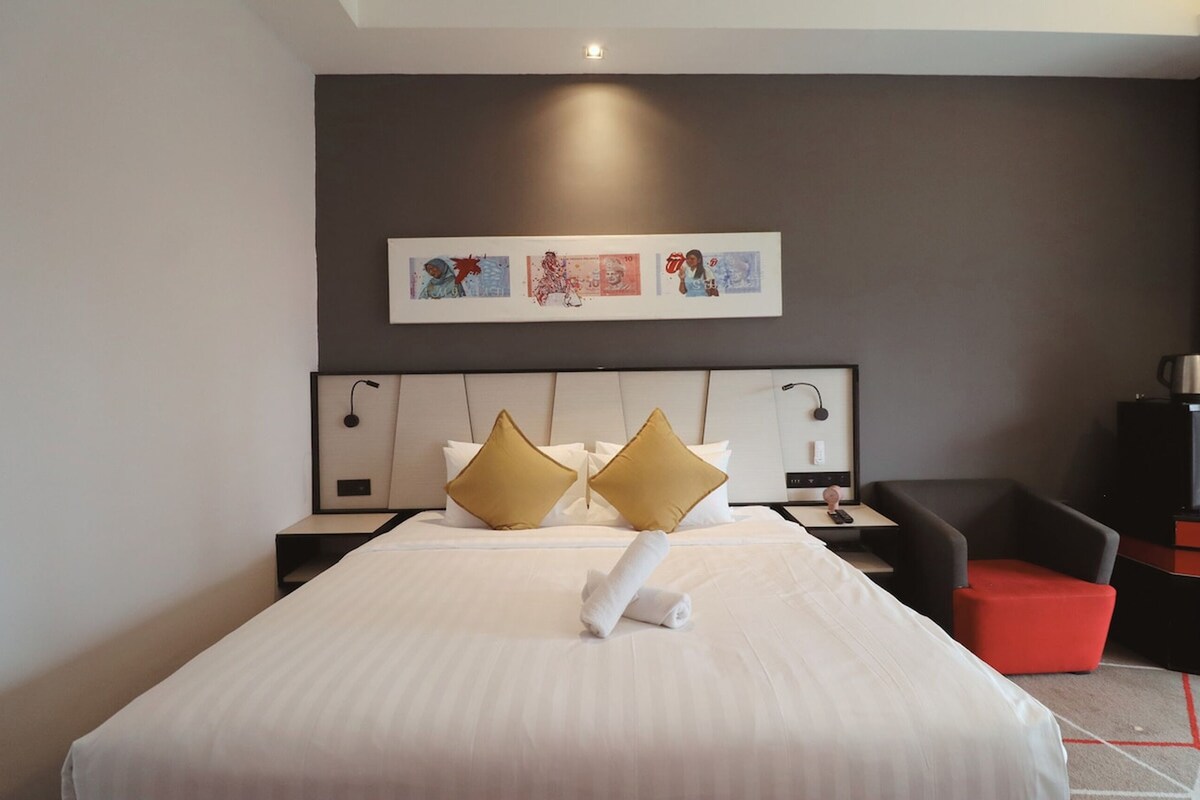 Una habitación de hotel con una cama grande, dos almohadas doradas, una colcha blanca, un reposapiés rojo, una silla negra y una obra de arte enmarcada en la pared.