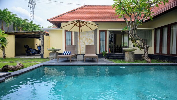 A swimming pool with lounge chairs and an umbrella, surrounded by a house with a red roof and large windows.
