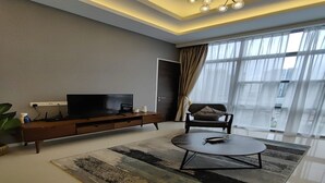 Een moderne woonkamer met een flatscreen televisie op een houten dressoir, een ronde salontafel en een groot raam met gordijnen.