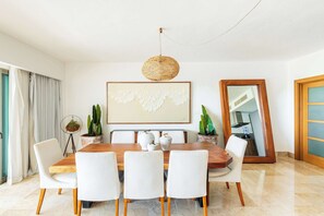 Uma sala de jantar com uma mesa de madeira, cadeiras brancas, um lustre pendurado, uma grande obra de arte emoldurada e um espelho na parede.