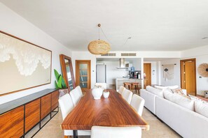Uma sala de estar moderna com uma mesa de jantar de madeira, sofás brancos e uma cozinha ao fundo.