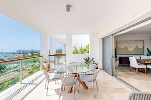 Varanda com mesa e cadeiras de vidro, vista para o mar e área de refeições com mesa e cadeiras.