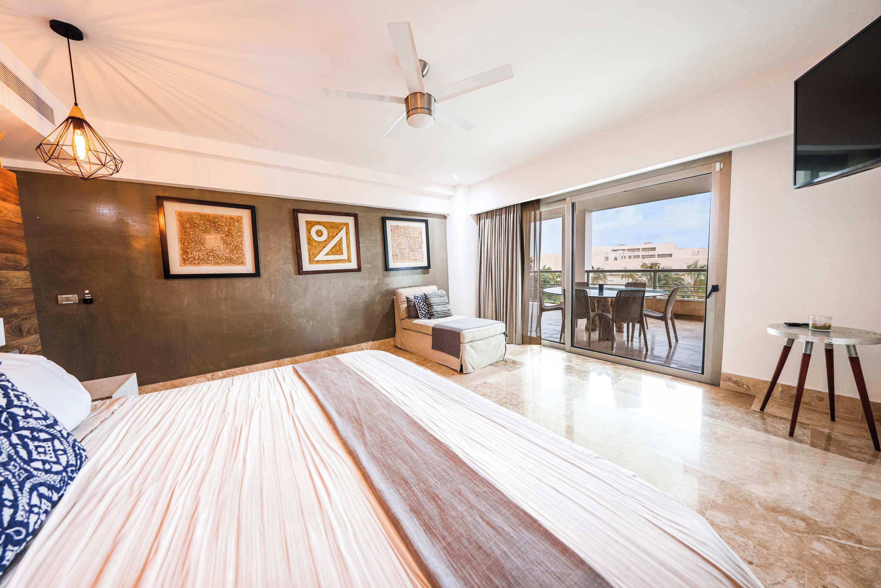 Ein modernes Schlafzimmer mit einem großen Bett, einem Deckenventilator, einem Balkon mit Tisch und Stühlen sowie gerahmten Kunstwerken an der Wand.