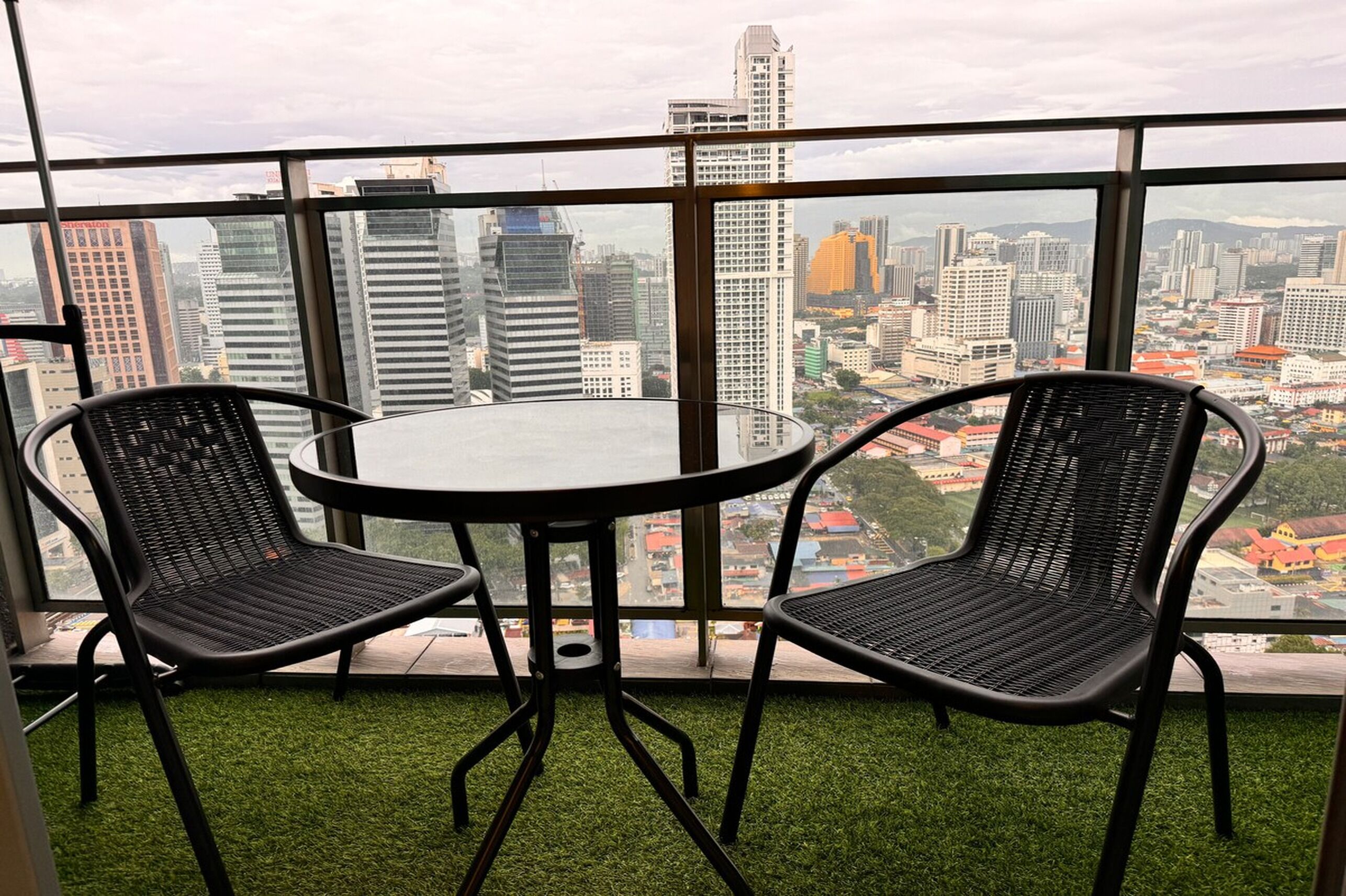 Ein Balkon mit zwei schwarzen Korbstühlen und einem runden Tisch mit Blick auf eine Stadtlandschaft und verschiedenen Gebäuden.