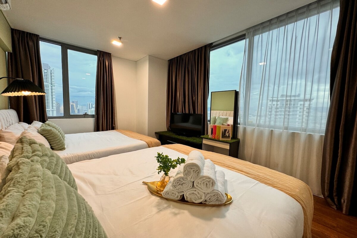 A hotel room with two beds, a TV, a view of the city, and a tray of towels.