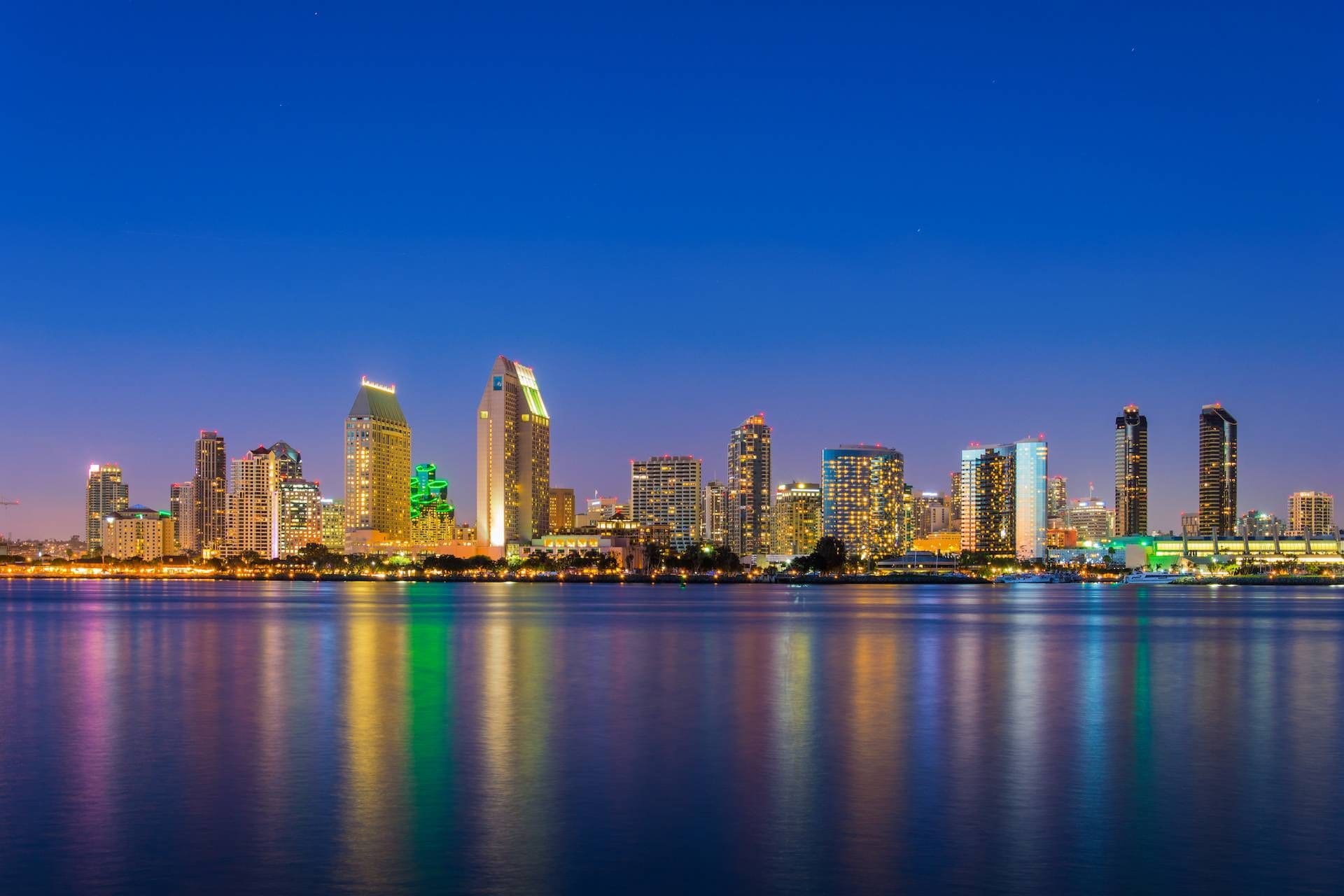 Silhueta urbana ao entardecer, com edifícios iluminados refletindo na água.