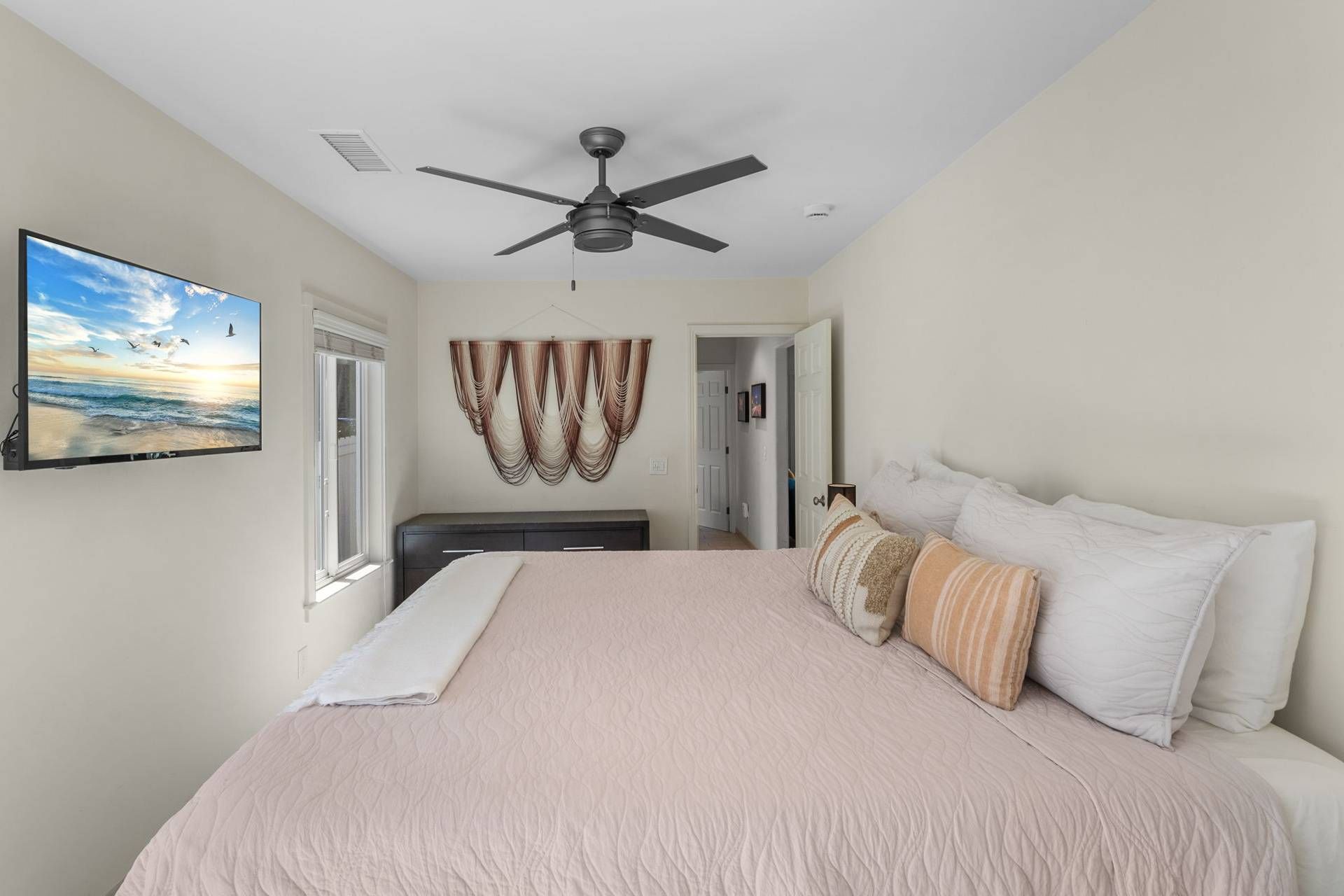 A bedroom with a bed, a ceiling fan, a TV on the wall, and a view of the outdoors.