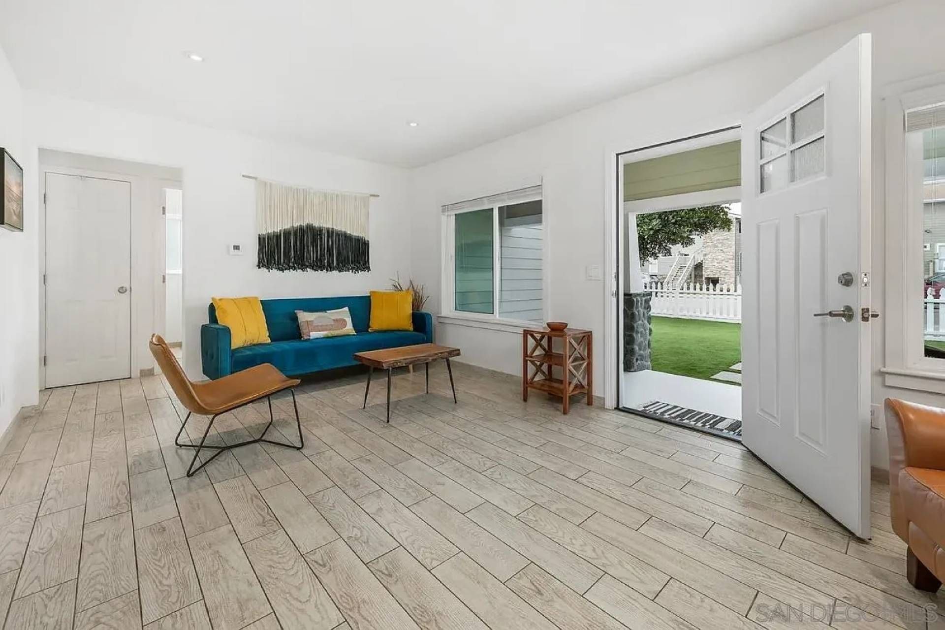 A modern living room with a blue sofa, a brown chair, and a small table. There is a door leading to a balcony with a view of a white fence and greenery.