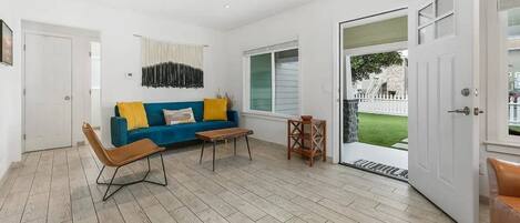 A modern living room with a blue sofa, a brown chair, and a small table. There is a door leading to a balcony with a view of a white fence and greenery.