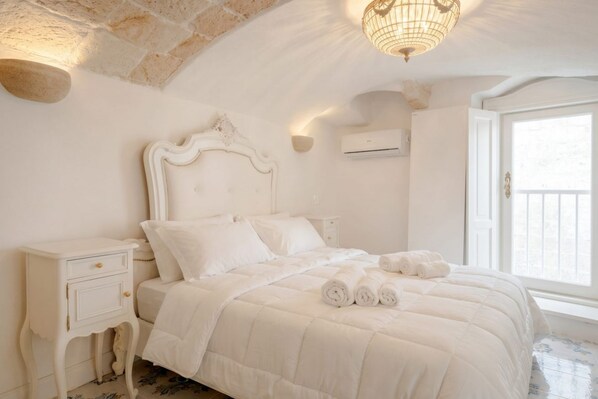Dormitorio con decoración blanca que incluye una cama, una mesita de noche, un aire acondicionado de pared y una ventana.