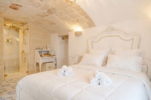 Habitación de hotel con una cama grande, zona de ducha, una mesita con artículos de aseo y cabecera blanca.