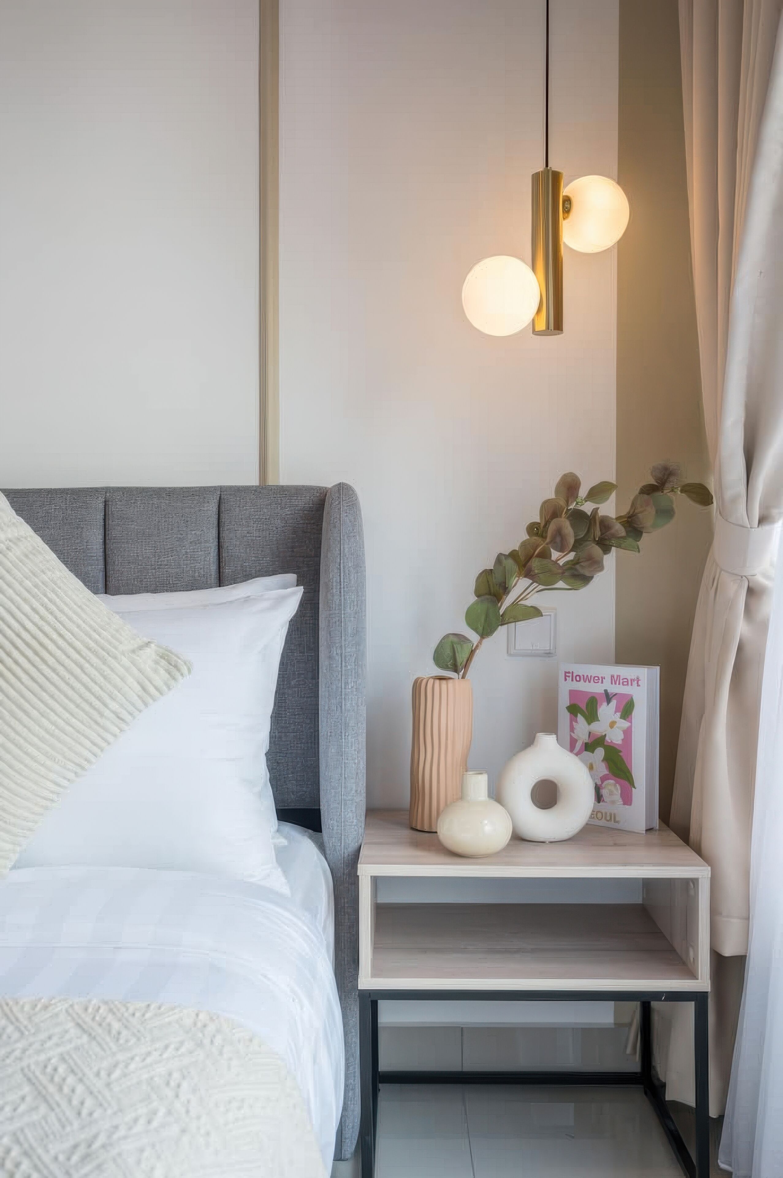 A neatly arranged bedroom with a grey upholstered headboard, a white bedspread, and a bedside table with a vase, a book titled 'Flower Mart', and a small white lamp.