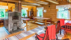 Interior rústico de una cabaña de madera con chimenea de piedra, mesa de comedor de madera, bancos y una araña.