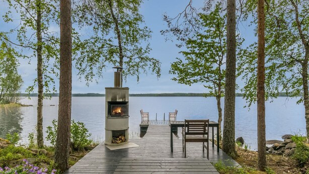 Una terraza de madera con una chimenea de piedra, dos sillas y una mesa con vista a un lago.