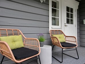 Deux fauteuils en osier avec des coussins, sur une véranda avec une porte blanche et un pot de fleurs.