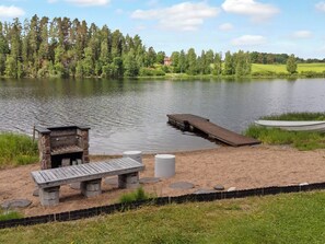 Un entorno junto al lago con un muelle de madera, una parrilla de piedra y una mesa de picnic.