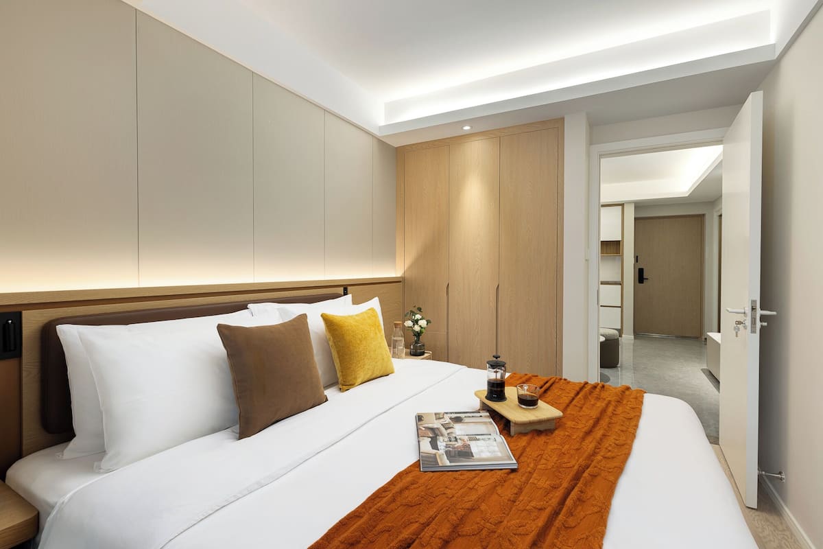 Quarto de hotel moderno com uma cama grande, um guarda-roupa de madeira, uma mesa de cabeceira com uma revista e um copo de água, e vista para o corredor.