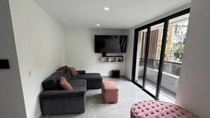 Un salon moderne avec un canapé d’angle gris, un pouf rose et un téléviseur à écran plat fixé au mur.