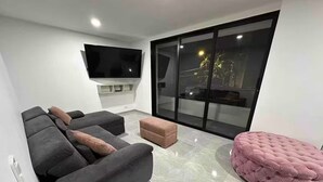Un salon moderne avec un grand téléviseur à écran plat fixé au mur, un canapé d’angle gris avec des coussins, un pouf rose et une porte-fenêtre coulissante donnant sur un espace extérieur.