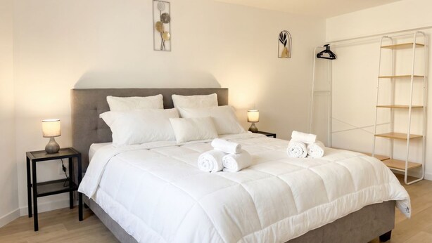 Una cama bien hecha con ropa blanca, dos mesitas de noche con lámparas y un armario blanco con estantes.