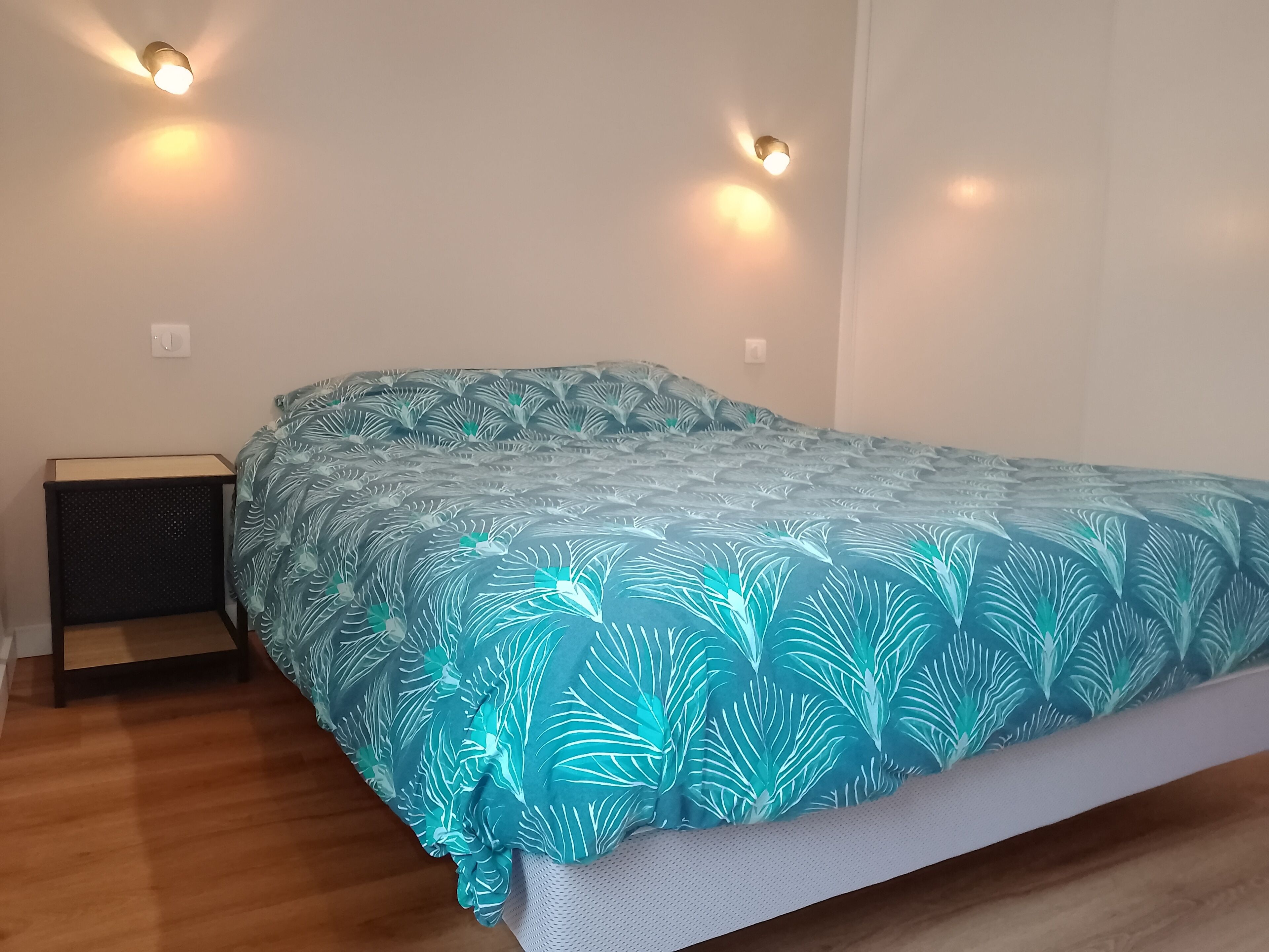 Una cama con una colcha estampada, dos lámparas de pared, una mesita de noche y un enchufe.
