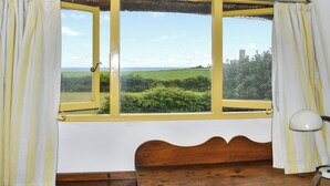Una habitación con una cama de madera, un ventanal de marcos amarillos y vistas a campos verdes y un edificio lejano.