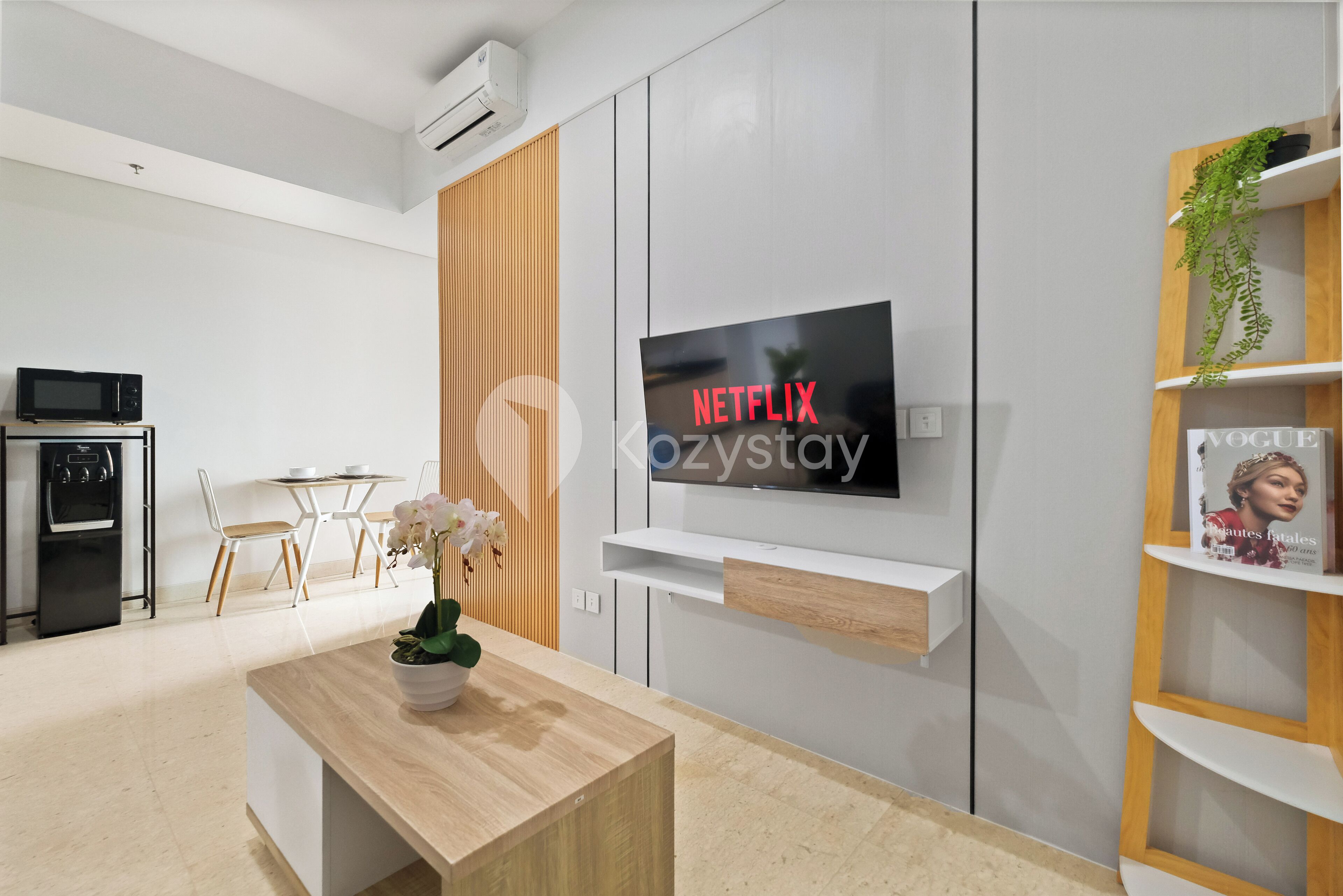 Uma sala de estar moderna com uma TV de tela plana na parede, uma mesa de centro de madeira, uma área de jantar com mesa e cadeiras brancas, e uma cozinha compacta com micro-ondas e forno.