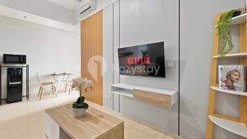 Uma sala de estar moderna com uma TV de tela plana na parede, uma mesa de centro de madeira, uma área de jantar com mesa e cadeiras brancas, e uma cozinha compacta com micro-ondas e forno.