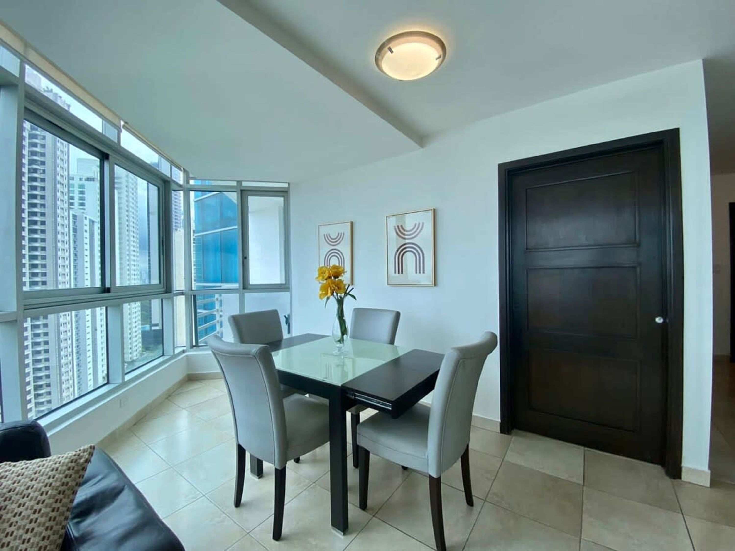 A modern dining area with a glass table, four chairs, a vase with yellow flowers, and a dark wooden door.