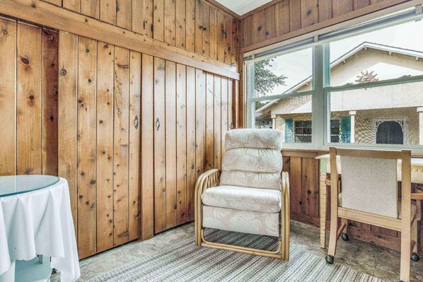 A room with wooden paneling, a cushioned chair, a small table with a white tablecloth, and a view through a window. - Adorable Navy Point Cottage - Pet Friendly - Conveniently Located - Watch The Blue Angels Practice (Pensacola)