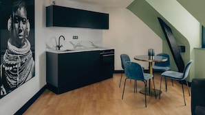 A modern kitchen with a black countertop, white backsplash, and a built-in dishwasher. There is a round table with blue chairs and a black sofa in the dining area. - Nemours Hotel (Rennes)