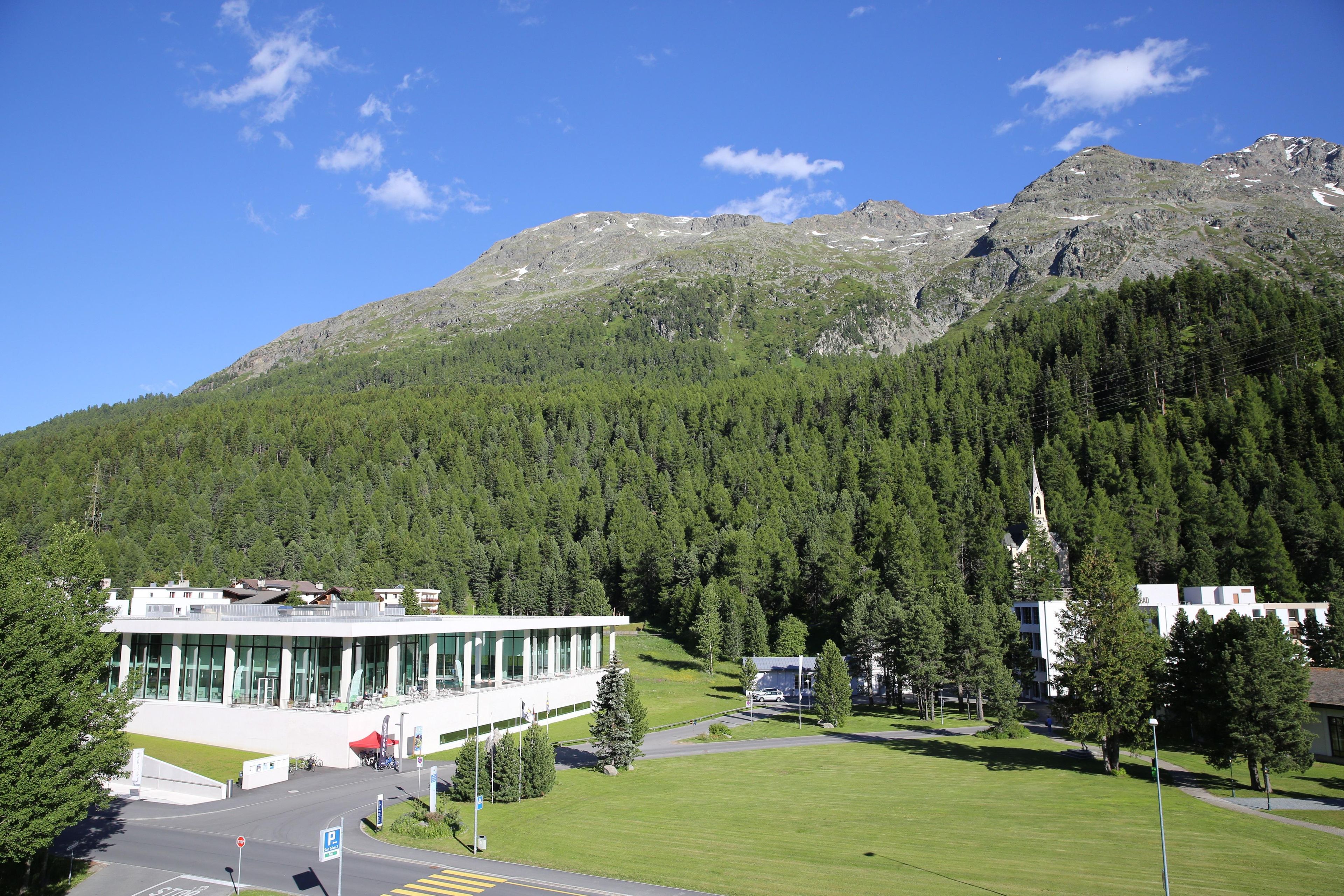 A modern building with a glass facade, surrounded by a well-maintained lawn and a forested mountain backdrop.
