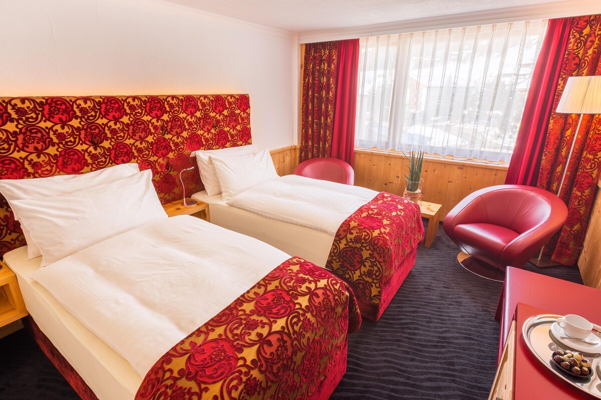 A hotel room with two beds, a red chair, a small table with a tray of snacks, and a window with curtains.