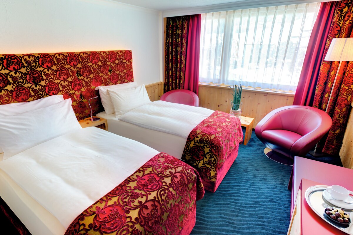 A hotel room with two beds, a red chair, a small table, and a window with curtains.