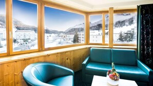 A room with a large window offering a mountain view, two teal chairs, and a small table with a fruit bowl. - TOP Hotel San Gian St. Moritz (St. Moritz)