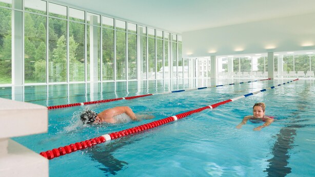 An indoor swimming pool with clear glass walls, red and white lane dividers, and a view of greenery outside. - TOP Hotel San Gian St. Moritz (St. Moritz)