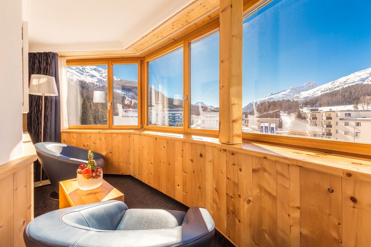 A room with a wooden interior, a large window with a mountain view, a massage chair, a small table with a fruit bowl, and a lamp.