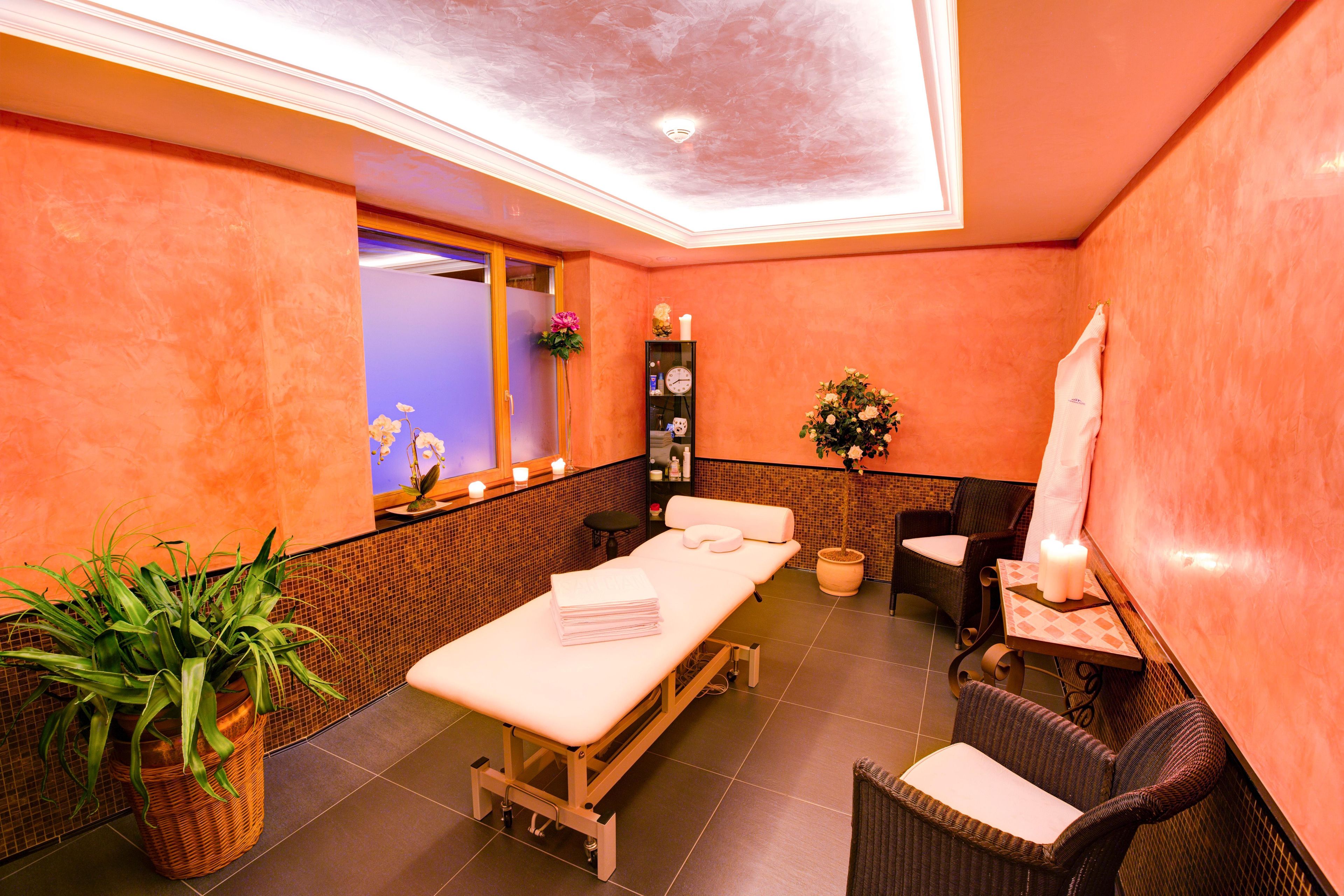 Ein Spa-Raum mit Massageliege, Stühlen, einer Topfpflanze und einem Regal mit Kerzen.