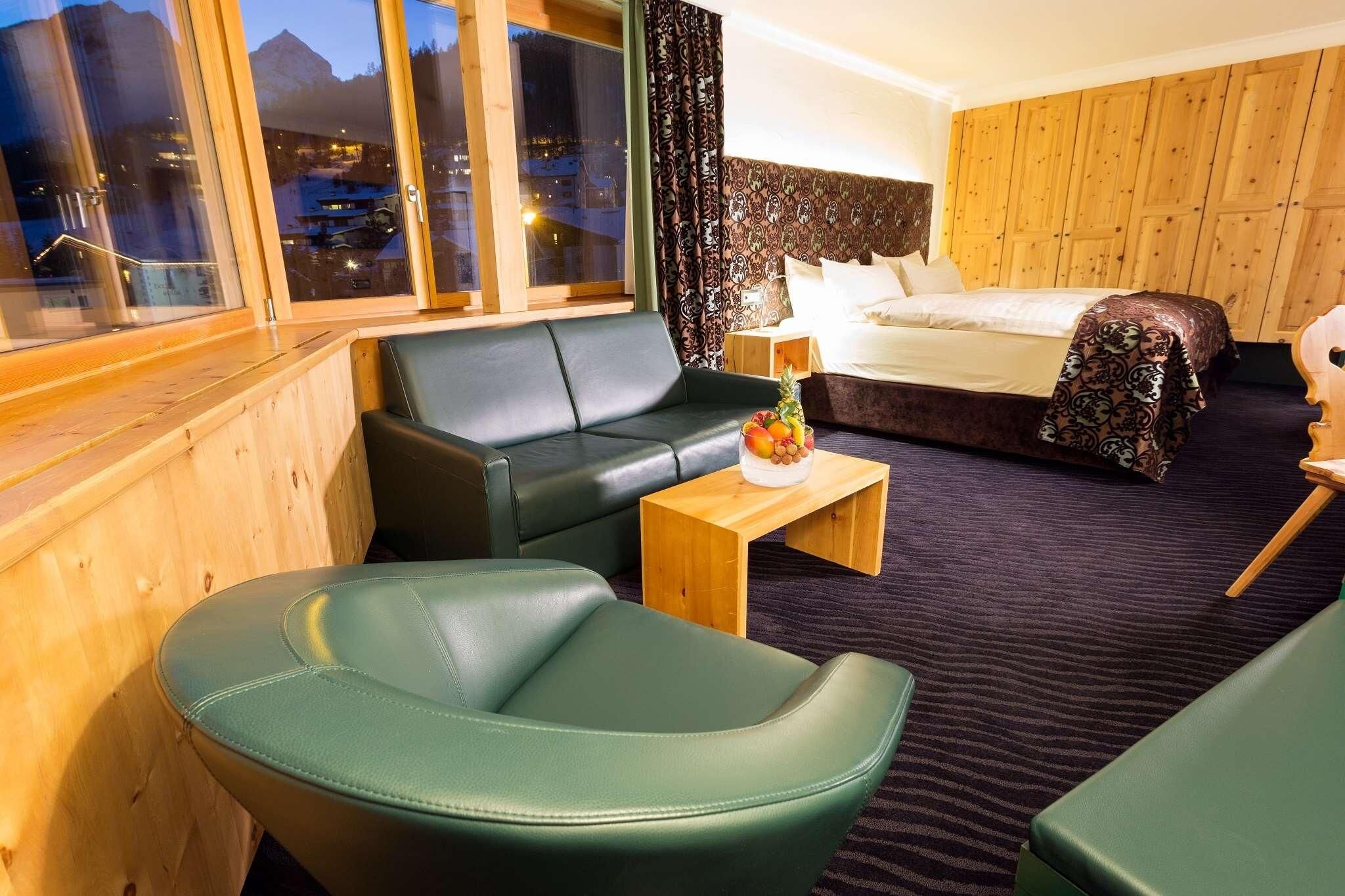 A hotel room with a bed, a green sofa, a wooden coffee table with fruit, and a view of mountains through large windows.