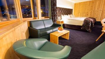 A hotel room with a bed, a green sofa, a wooden coffee table with fruit, and a view of mountains through large windows.