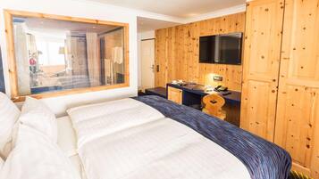 A hotel room with a large bed, a wooden wardrobe, a television mounted on the wall, and a small desk with a telephone.