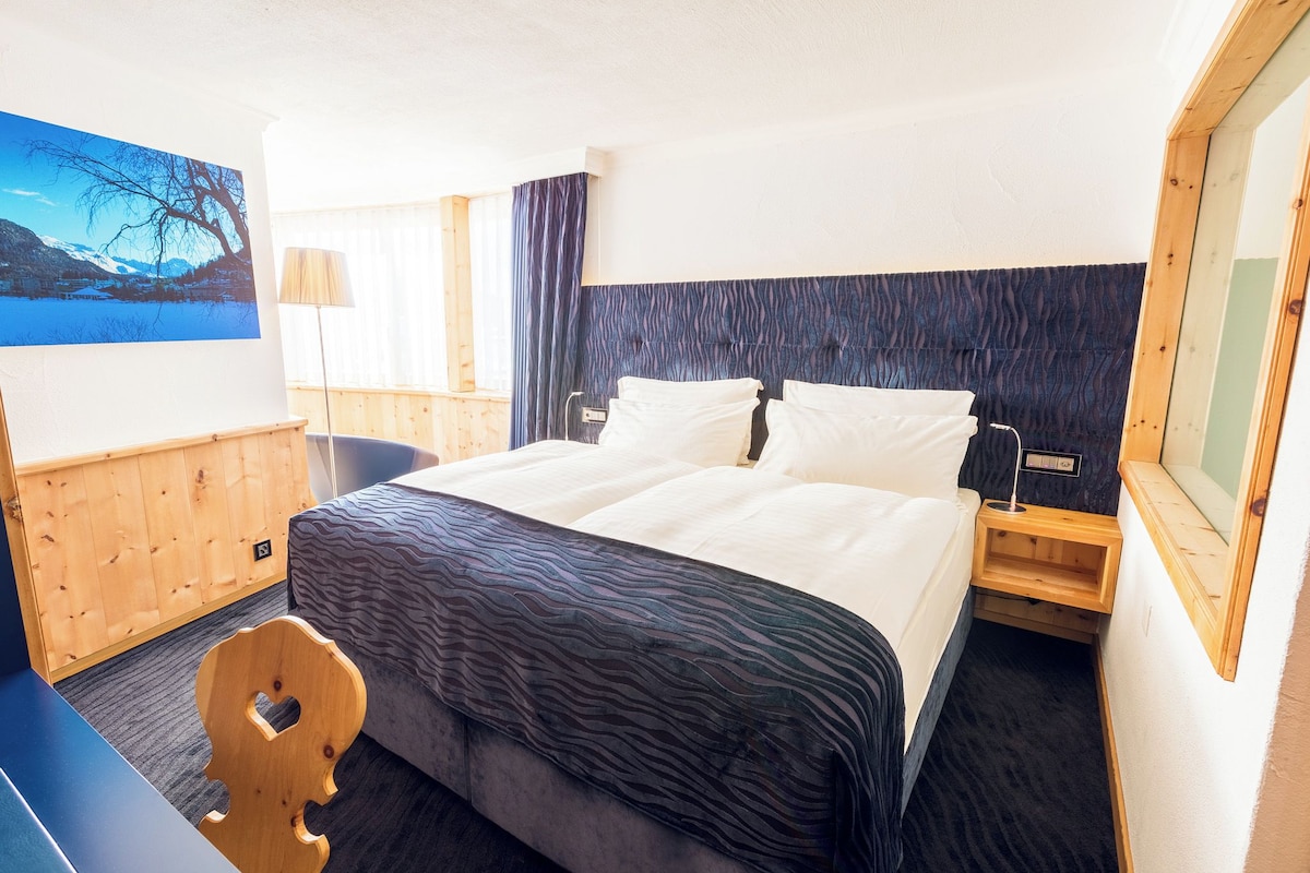 A hotel room with a bed, a desk, a chair, a lamp, and a painting of a landscape.