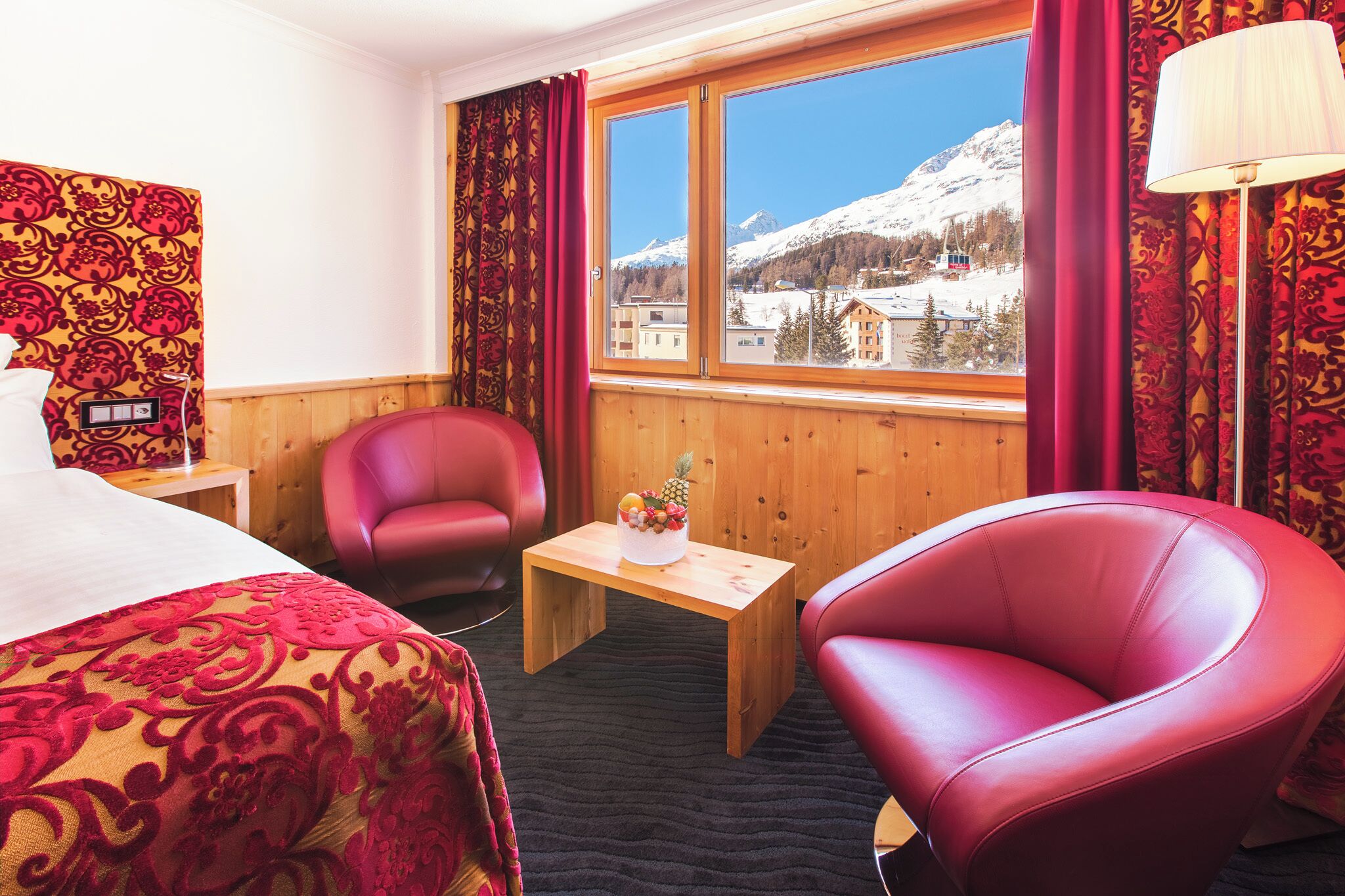 A hotel room with a bed, a wooden table, a red chair, and a view of snow-covered mountains.